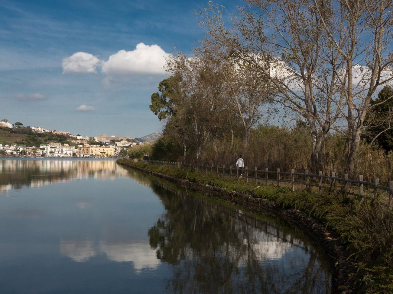 Le acque calme del Lago d'Averno che riflettono il cielo, con un sentiero lungo la riva e un paese dei Campi Flegrei in lontananza