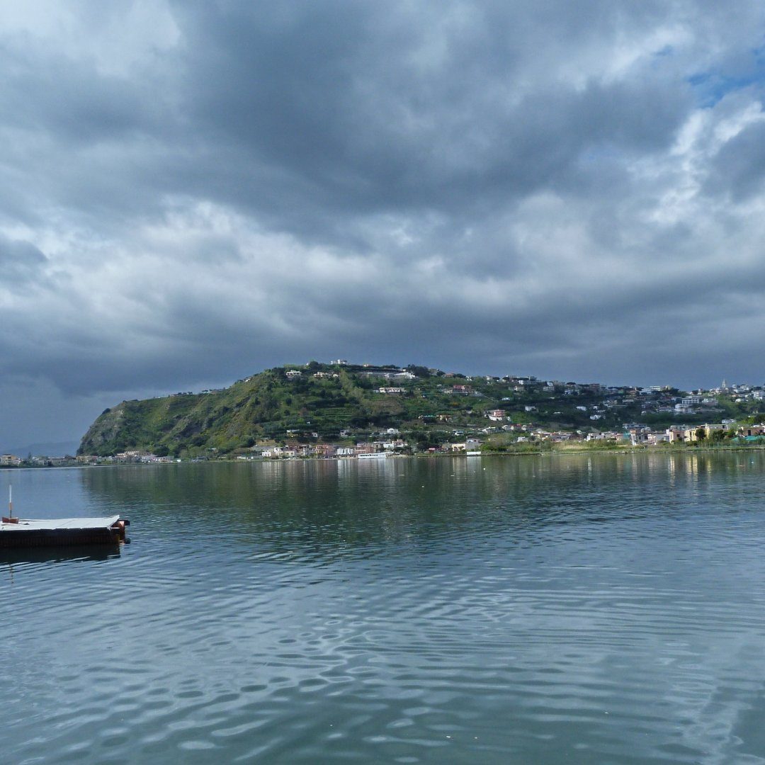 Le acque verdi e calme del Lago di Miseno a Bacoli sotto un cielo nuvoloso e drammatico, con il paese sulla collina sullo sfondo